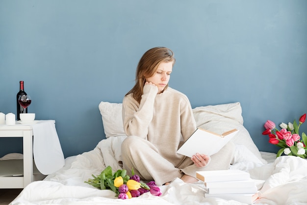 Heureuse femme souriante assise sur le lit en pyjama, avec plaisir en appréciant les fleurs et en lisant un livre