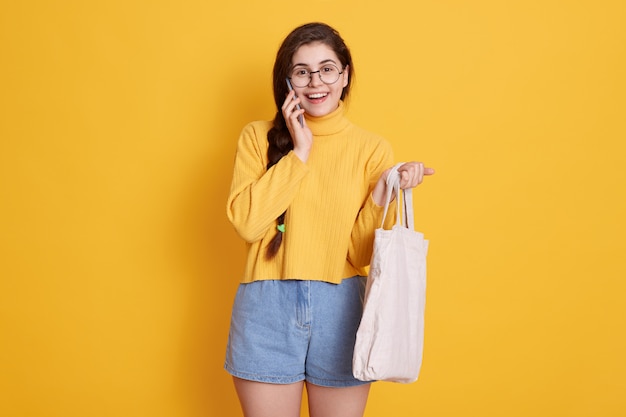 Heureuse femme souriante aux cheveux noirs et queue de cochon posant contre le mur jaune et parler par téléphone, tenant un sac en coton dans les mains, portant un pull et un jean court.