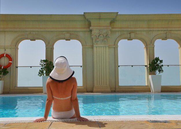 Heureuse femme souriante avec chapeau et lunettes de soleil dans la piscine du complexe tropical