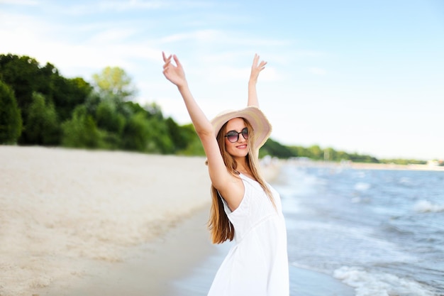 Heureuse femme souriante dans le bonheur du bonheur gratuit sur la plage de l'océan, debout avec un chapeau, des lunettes de soleil et des mains rasées. Portrait d'un modèle féminin multiculturel en robe d'été blanche profitant de la nature pendant les voyages