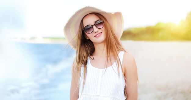 Heureuse femme souriante dans le bonheur gratuit sur la plage de l'océan debout avec un chapeau, des lunettes de soleil et des fleurs blanches. Portrait d'un modèle féminin multiculturel en robe d'été blanche profitant de la nature