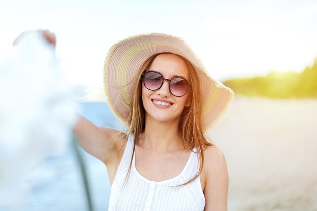 Heureuse femme souriante dans le bonheur gratuit sur la plage de l'océan debout avec un chapeau, des lunettes de soleil et des fleurs blanches. Portrait d'un modèle féminin multiculturel en robe d'été blanche profitant de la nature