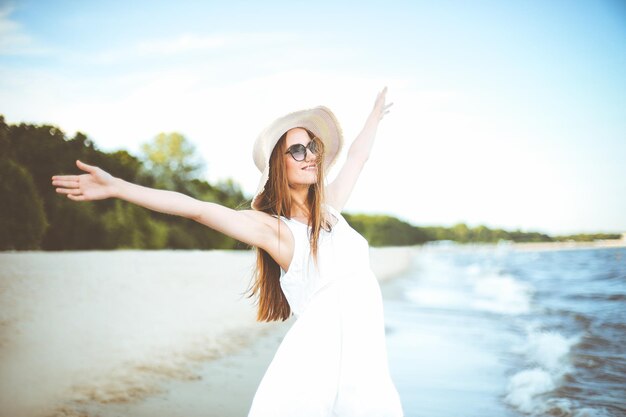 Heureuse femme souriante dans le bonheur gratuit sur la plage de l'océan, debout avec un chapeau, des lunettes de soleil et des mains rasantes. Portrait d'un modèle féminin multiculturel en robe d'été blanche profitant de la nature pendant le voyage