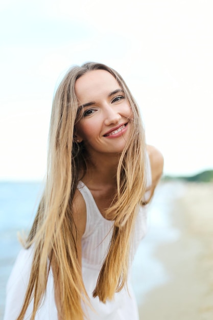Heureuse femme souriante dans le bonheur gratuit sur la plage de l'océan en regardant la caméra. Portrait d'un modèle féminin multiculturel en robe d'été blanche profitant de la nature pendant les vacances de voyage en plein air