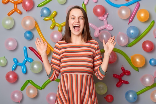 Heureuse femme souriante excitée vêtue d'une robe rayée debout contre un mur gris avec décoration de ballons bras levés criant d'étonnement célébrant l'ambiance festive