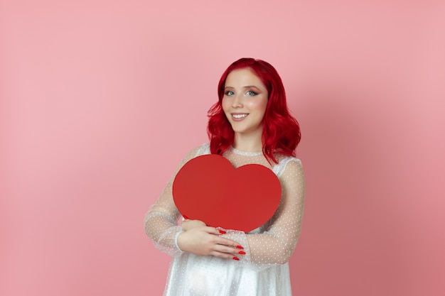 Heureuse femme souriante en robe blanche et aux cheveux rouges tenant un grand coeur de papier rouge