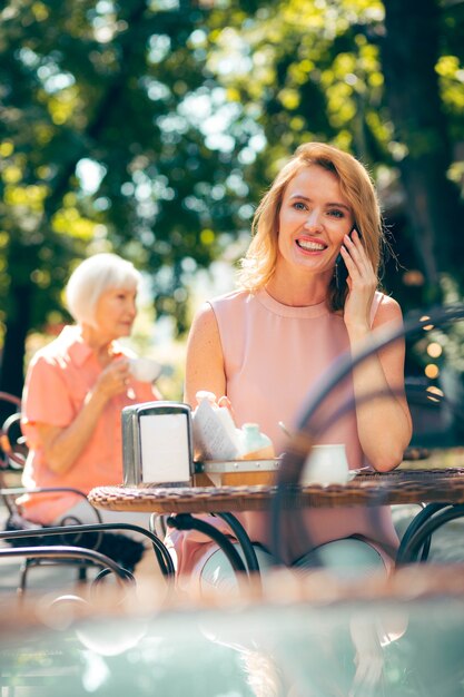 Heureuse femme souriante à la table du café à l'extérieur, profitant d'une agréable conversation téléphonique