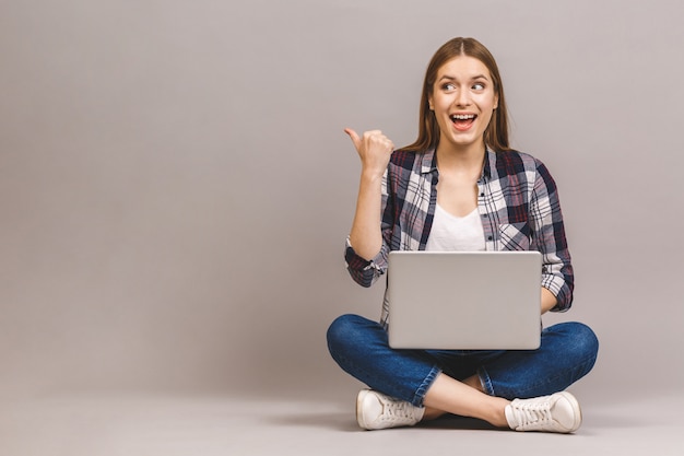 Heureuse femme souriante travaillant sur un ordinateur portable assis sur le sol avec les jambes croisées et pointant le doigt loin