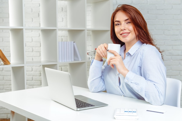 Heureuse femme souriante travaillant avec un ordinateur portable et de boire du café