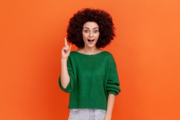 Heureuse femme surprise avec une coiffure Afro portant un chandail décontracté vert levant le doigt, a une bonne idée soudaine, regardant la caméra avec la bouche ouverte. Studio intérieur tourné isolé sur fond orange.