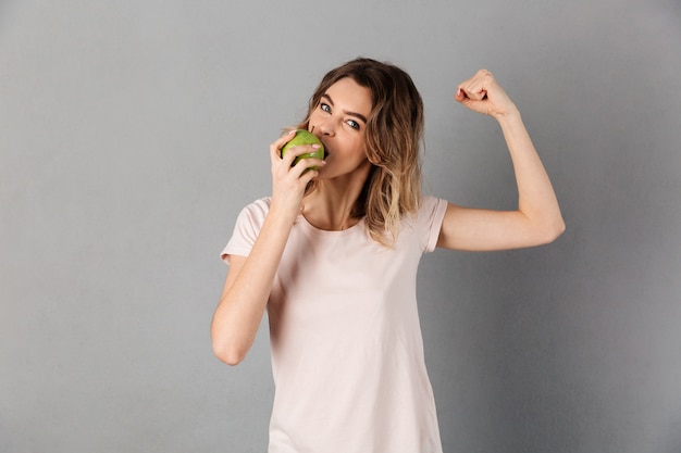 Heureuse femme en t-shirt mange une pomme et montre son biceps tout en gris