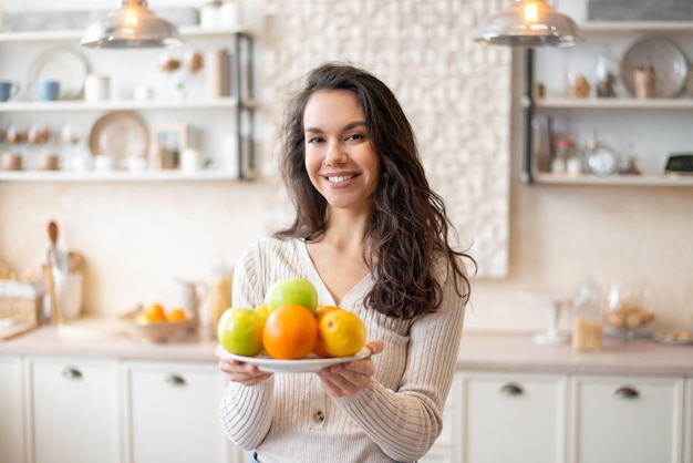 Heureuse femme tenant une assiette avec des fruits frais regardant et souriant à la caméra debout dans la cuisine