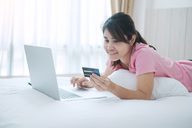 Heureuse femme tenant une carte de crédit et utilisant un ordinateur portable pour les achats en ligne tout en passant des commandes sur le lit le matin à la maison.