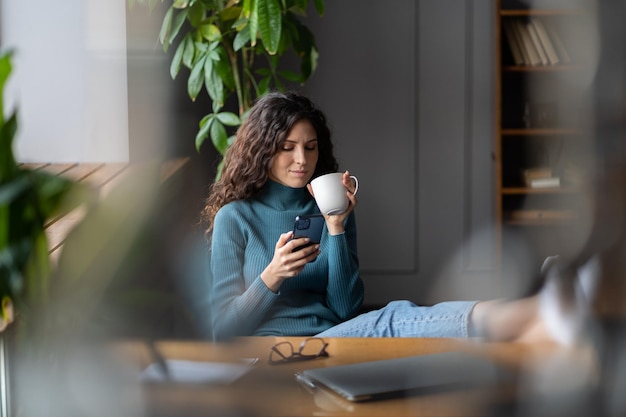 Heureuse femme tergiversant sur le lieu de travail, boire du café, lire les actualités en ligne sur un smartphone au bureau