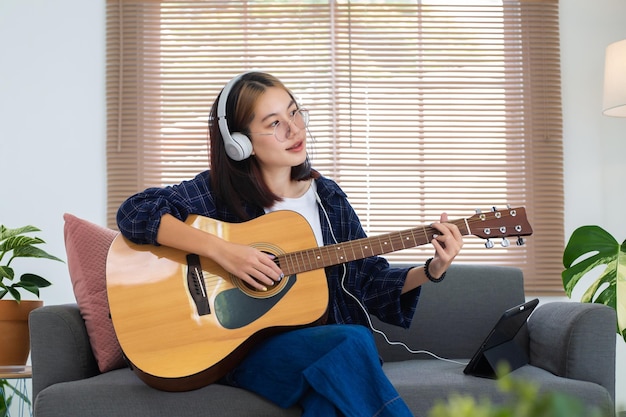 Heureuse fille asiatique à lunettes jouant de la guitare acoustique et écoutant de la musique à partir d'une tablette dans le salon à la maison Concept de loisirs à la maison