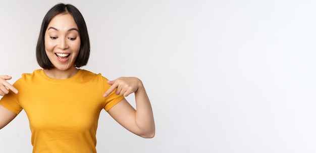 Heureuse fille asiatique pointant les doigts vers le bas en regardant la bannière d'annonce ou la publicité debout en t-shirt jaune sur fond blanc