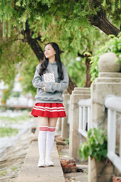 Heureuse fille asiatique en uniforme scolaire de sweat à capuche, jupe et chaussettes hautes serrées debout à l'extérieur avec un livre dans les mains