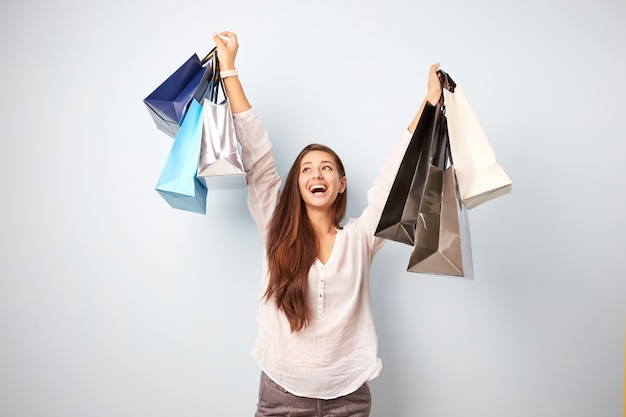 Heureuse fille aux cheveux bruns vêtue d'un chemisier blanc et d'un pantalon gris se dresse avec beaucoup de sacs après avoir fait du shopping sur fond blanc.