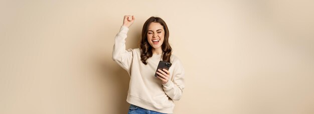 Heureuse fille brune tenant un smartphone et acclamant la victoire en célébrant la victoire sur l'application de téléphone mobile stan