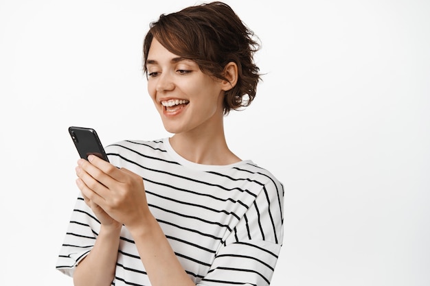 Heureuse fille brune utilisant un téléphone portable, souriant et regardant le téléphone, debout en t-shirt rayé sur blanc