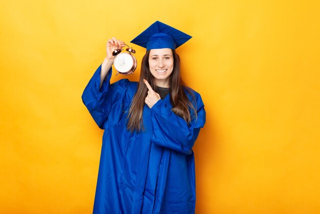 Heureuse fille diplômée pointe vers le réveil, disant son heure.