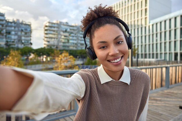 Heureuse fille du millénaire optimiste avec hiar bouclé écoute de la musique via des écouteurs sans fil étend le bras avec un appareil inconnu pose pour faire des selfies porte des vêtements soignés profite du beau temps pendant la journée