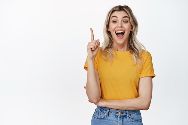 Heureuse fille excitée pointant le doigt vers le haut en riant et en souriant montrant le logo ou la bannière au-dessus debout sur fond isolé blanc