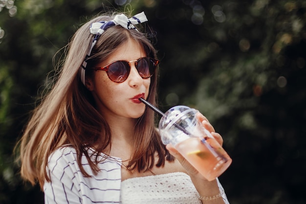 Heureuse fille hipster avec des lunettes de soleil en robe rétro et bandeau tenant une tasse en plastique et de la paille buvant de la limonade Belle jeune femme élégante tenant un verre et se détendre dans la rue de la ville