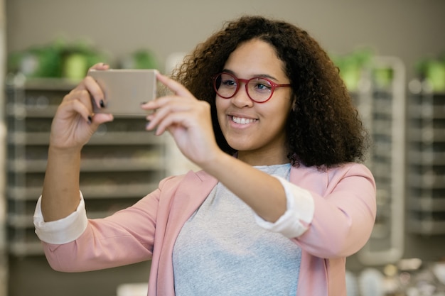 Heureuse fille interculturelle à lunettes tenant le smartphone devant son visage tout en essayant de nouvelles lunettes et en faisant selfie