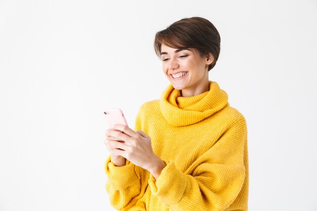 Heureuse fille joyeuse portant un sweat à capuche debout isolé sur blanc, à l'aide de téléphone mobile