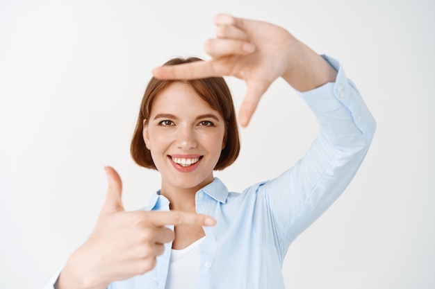 Heureuse fille naturelle faisant une photo de cadre à la main et souriante, capturant le moment, planifiant une position parfaite, debout sur un mur blanc