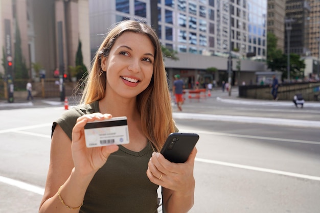 Heureuse fille rêveuse regardant ailleurs décider ce qu'elle achète à l'aide d'un smartphone et montrant une carte de crédit dans la rue de la métropole