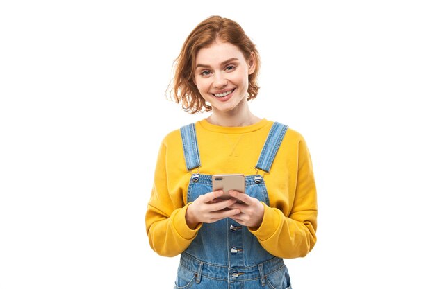 Heureuse fille rousse souriante tenant un smartphone dans les mains dans des vêtements jaunes isolés sur fond de studio blanc, a reçu un message joyeux