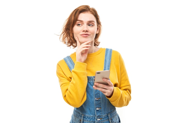 Heureuse fille rousse souriante tenant un smartphone dans les mains dans des vêtements jaunes isolés sur fond de studio blanc, a reçu un message joyeux