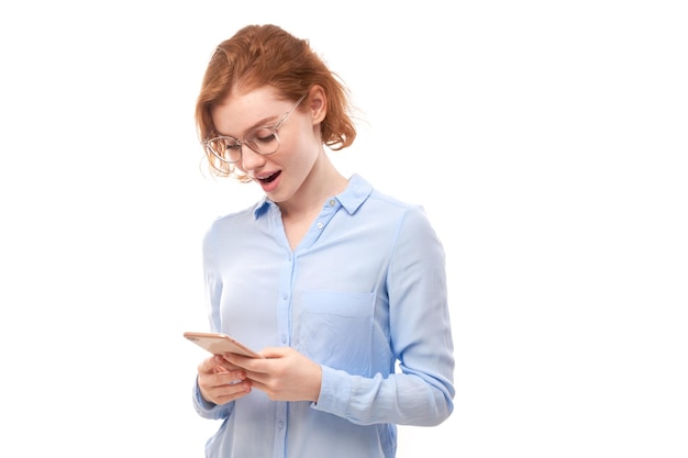 Heureuse fille rousse souriante tenant un smartphone dans les mains en lunettes et chemise d'affaires isolée sur fond de studio blanc, a reçu un message joyeux