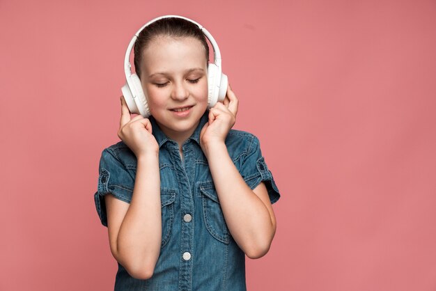 Heureuse fille satisfaite en jeans T-shirt debout et écoutant sa musique préférée avec des écouteurs, les yeux fermés et souriant. Prise de vue en studio intérieur, isolé sur fond rose