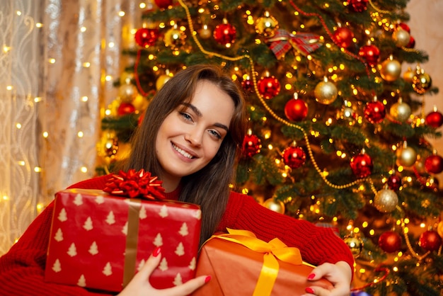 Heureuse fille souriante assise près d'un sapin de Noël décoré avec des boîtes à cadeaux dans ses mains Heureuse période magique des vacances d'hiver et atmosphère chaleureuse