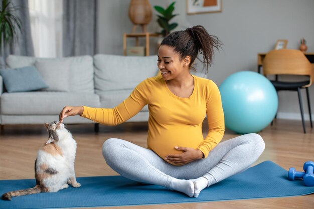Heureuse heureuse jeune femme afro-américaine enceinte avec un gros ventre pratique l'intérieur du yoga et joue avec un chat