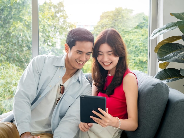 Heureuse jeune belle femme asiatique et bel homme sourient ensemble tout en regardant l'écran de la tablette sur un canapé gris près de la fenêtre en verre du salon blanc Couple amoureux utilisant une tablette numérique ensemble