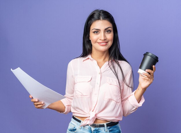 Heureuse jeune belle femme dans des vêtements décontractés tenant un document et une tasse de café à la recherche de sourire confiant