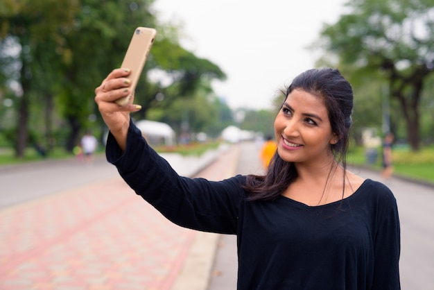 Heureuse jeune belle femme persane prenant selfie dans le parc