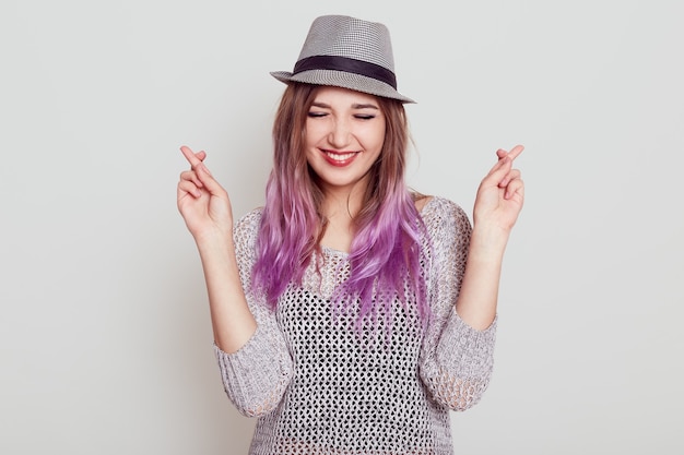 Heureuse jeune belle femme portant chemise et chapeau croisant les doigts, espère quelque chose de bien, garde les yeux fermés et a un sourire à pleines dents, isolé sur un mur blanc.