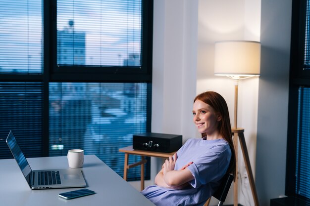 Heureuse jeune blogueuse rousse satisfaite d'avoir terminé l'article à temps en terminant un travail à distance tard dans la nuit. Femme journaliste souriante travaillant assise au bureau près de la fenêtre tard dans la soirée.