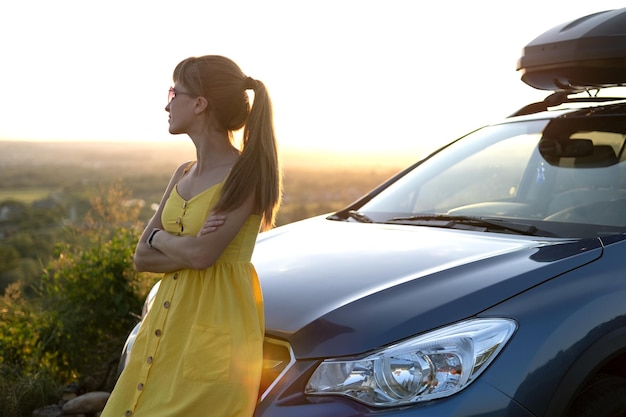 Heureuse jeune conductrice en robe jaune profitant d'une chaude soirée d'été appuyée sur sa voiture.