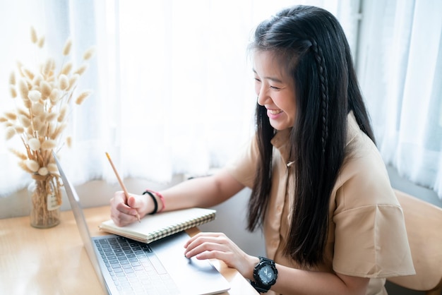 Photo heureuse de jeune étudiante adolescente asiatique prenant des notes écrites sur un bloc-notes occasionnel en utilisant un ordinateur portableconcept d'apprentissage électronique en lignepersonnes indépendantes femmes d'affaires