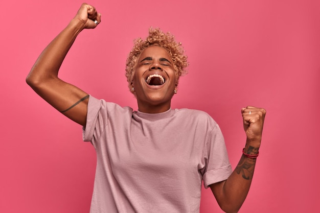 Heureuse jeune étudiante afro-américaine jeune fille se réjouit de lever les bras sur fond rose en studio Concept de gagner à la loterie et fan de votre équipe sportive préférée Espace publicitaire
