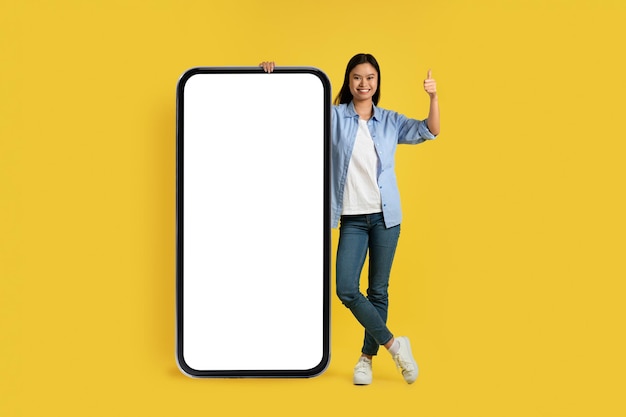 Heureuse jeune étudiante chinoise en spectacle décontracté pouce levé debout près d'un énorme smartphone avec écran vide