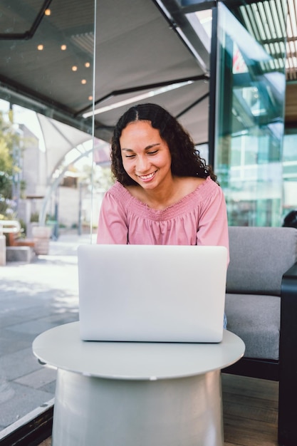 Heureuse jeune étudiante latine utilisant un ordinateur portable et assise à l'extérieur d'un café pendant son temps libre. Vertical