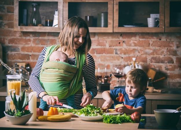 Heureuse jeune famille, belle mère de deux enfants, adorable garçon d'âge préscolaire et bébé en écharpe cuisiner ensemble dans une cuisine ensoleillée.