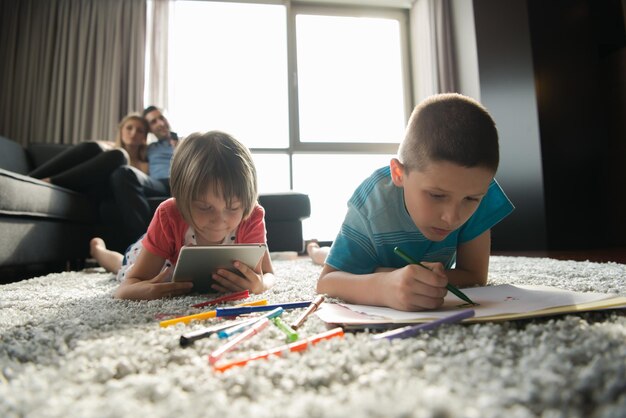 Heureuse jeune famille jouant ensemble à la maison sur le sol à l'aide d'une tablette et d'un ensemble de dessins pour enfants